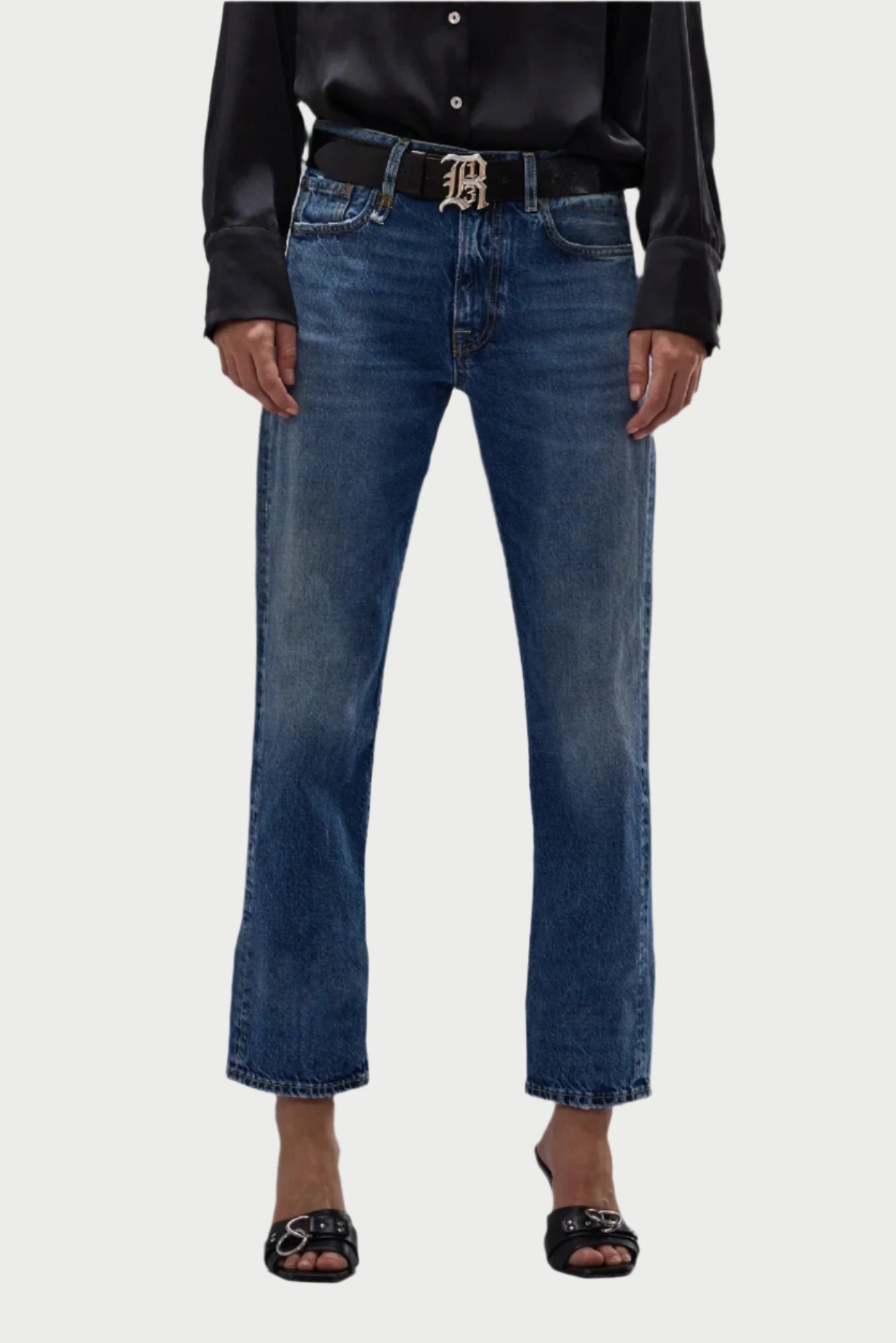 A person in an R13 Romeo Jean by R13, paired with a 100% cotton black shirt and black open-toed sandals, accessorized with a black belt featuring a decorative buckle. The plain light-colored background accentuates the elegance of this Made in Italy ensemble.