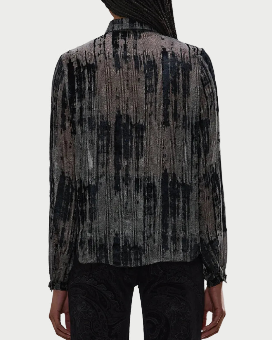 A person faces away, wearing the R13 Ruffle Blouse—a sheer grey top with a black abstract vertical pattern by R13. Only their upper body and part of the arms are visible.