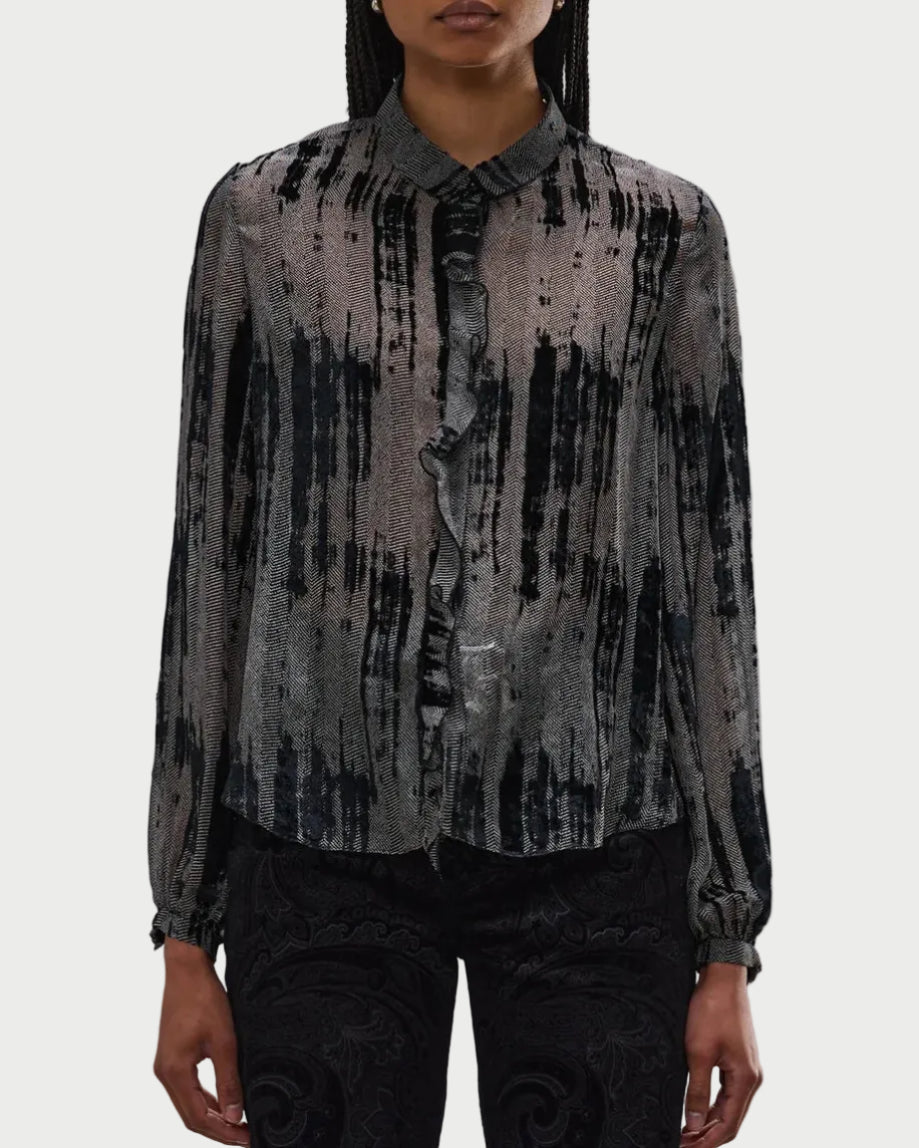 A person wears the R13 Ruffle Blouse by R13, featuring a black and gray abstract stripe pattern, front ruffle trim, and button-up closure, paired with dark patterned pants against a plain white background.