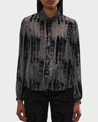 A person wears the R13 Ruffle Blouse by R13, featuring a black and gray abstract stripe pattern, front ruffle trim, and button-up closure, paired with dark patterned pants against a plain white background.