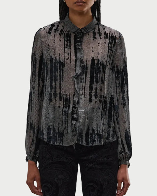 A person wears the R13 Ruffle Blouse by R13, featuring a black and gray abstract stripe pattern, front ruffle trim, and button-up closure, paired with dark patterned pants against a plain white background.