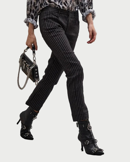 The lower half of a person is shown wearing the R13 Tailored Boy Straight Pant by R13 in black pinstripe, paired with black heeled ankle boots with buckles, a patterned long-sleeve top, and holding a small black chain-strap handbag.