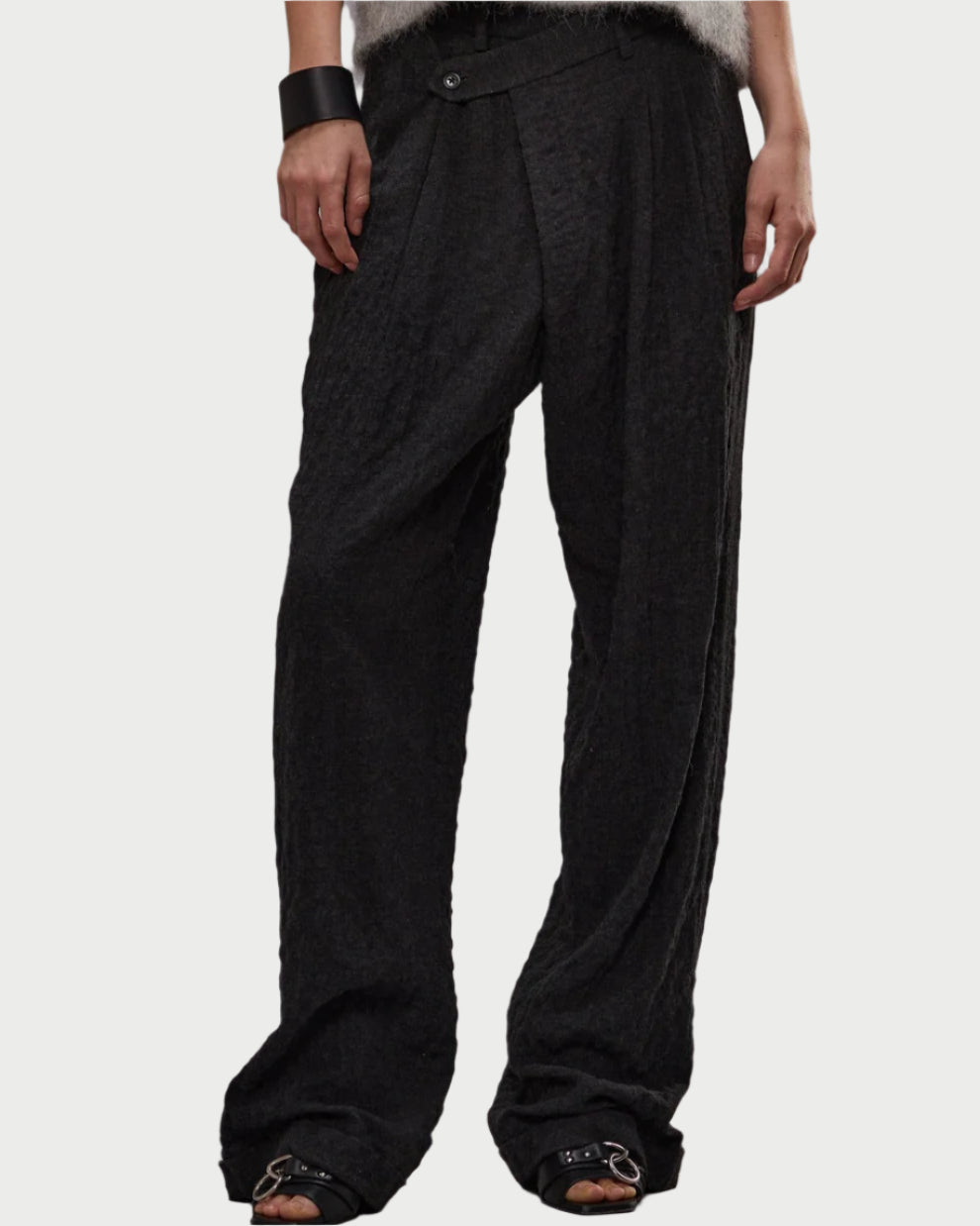 Someone wears loose-fitting, textured black R13 Triple Pleat Crossover Trousers by R13 in herringbone suiting with a slightly asymmetric waistband, paired with black open-toe sandals, a black wrist cuff, and a fuzzy white top; lower body is shown.