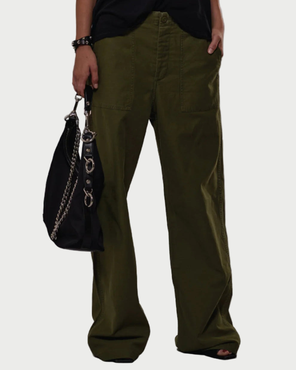 Person wearing olive green pants holding a black handbag against a gray background