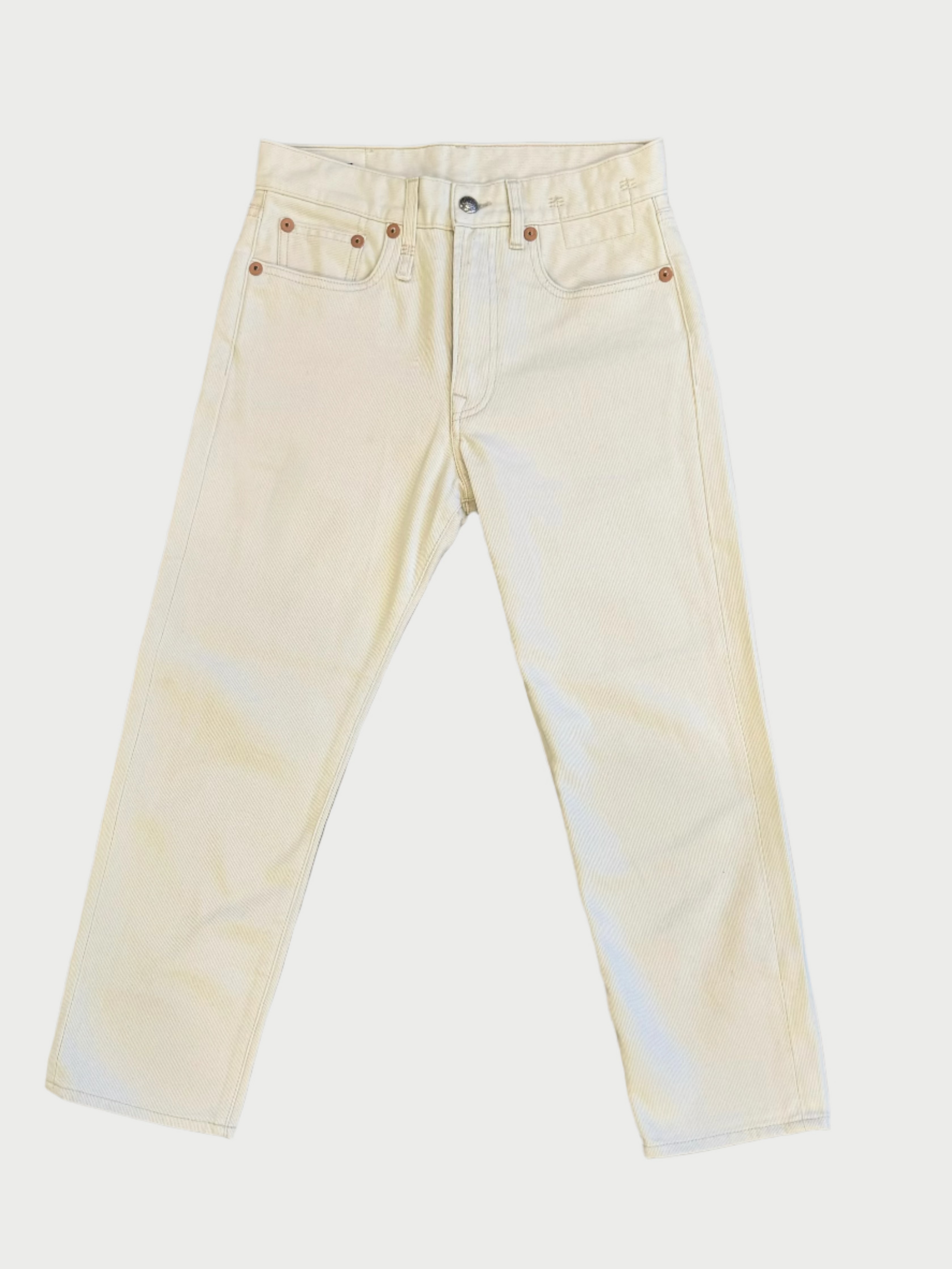A pair of cream-colored, straight-leg Corduroy Boyfriend Jean by R13 is laid out flat on a wooden surface. The jeans have a button closure, belt loops, and pockets with rivets near the top corners. The wooden surface has a light brown color with a visible grain pattern.