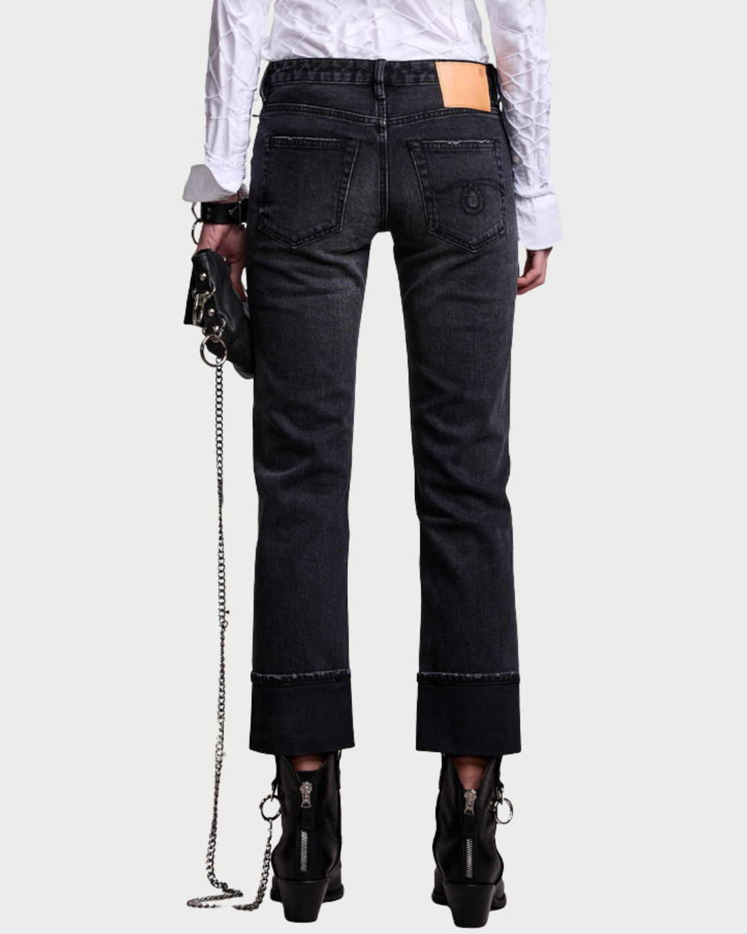 A person wearing the R13 Cuffed Boy Straight jeans—black wash straight-leg denim with rolled hems and a tan patch on the waistband—stands facing away. They pair the jeans with a white textured shirt, black heeled boots, and carry a small chain-strap bag in their left hand.