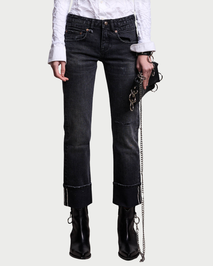 A person wearing a white textured shirt, the R13 Cuffed Boy Straight jeans in a black wash with visible seams and cuffed hems from R13, black heeled boots, and holding a small bag with a metal chain strap. The background is plain and light-colored.