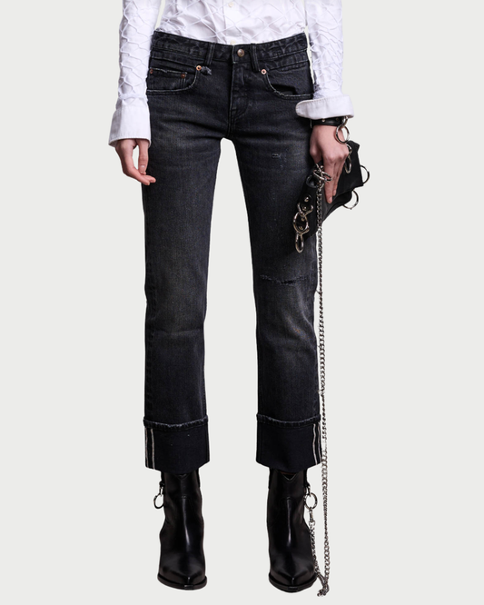 A person wearing a white textured shirt, the R13 Cuffed Boy Straight jeans in a black wash with visible seams and cuffed hems from R13, black heeled boots, and holding a small bag with a metal chain strap. The background is plain and light-colored.