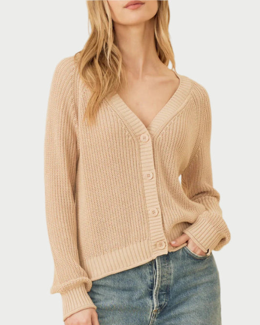 A person with long, wavy blonde hair is wearing the versatile One Grey Day Raleigh Cardigan in beige, featuring a V-neck and buttons down the front. They have paired it with blue jeans and are standing against a plain, light-colored background with a relaxed expression and one hand in their pocket.