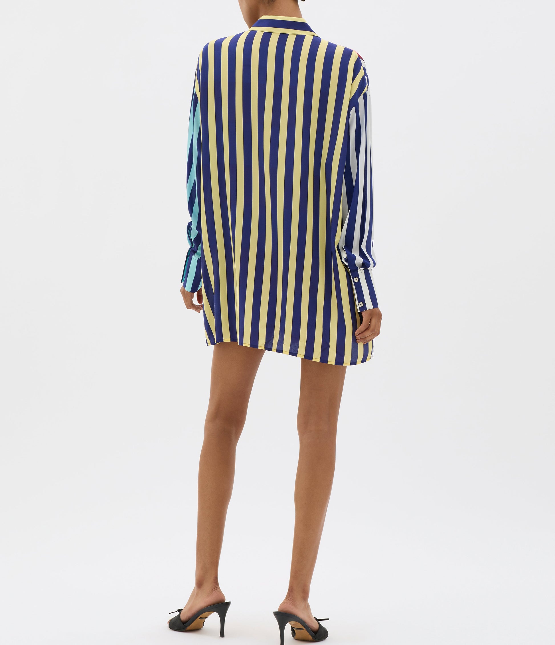 A person wears a MUMA Striped Oversized Shirt in vertical yellow, blue, and white stripes, mid-thigh length, paired with black heeled sandals against a plain white background.