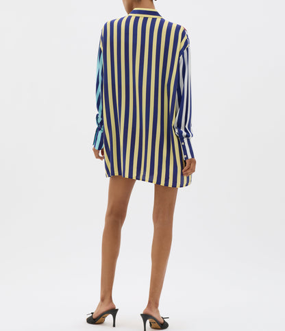 A person wears a MUMA Striped Oversized Shirt in vertical yellow, blue, and white stripes, mid-thigh length, paired with black heeled sandals against a plain white background.
