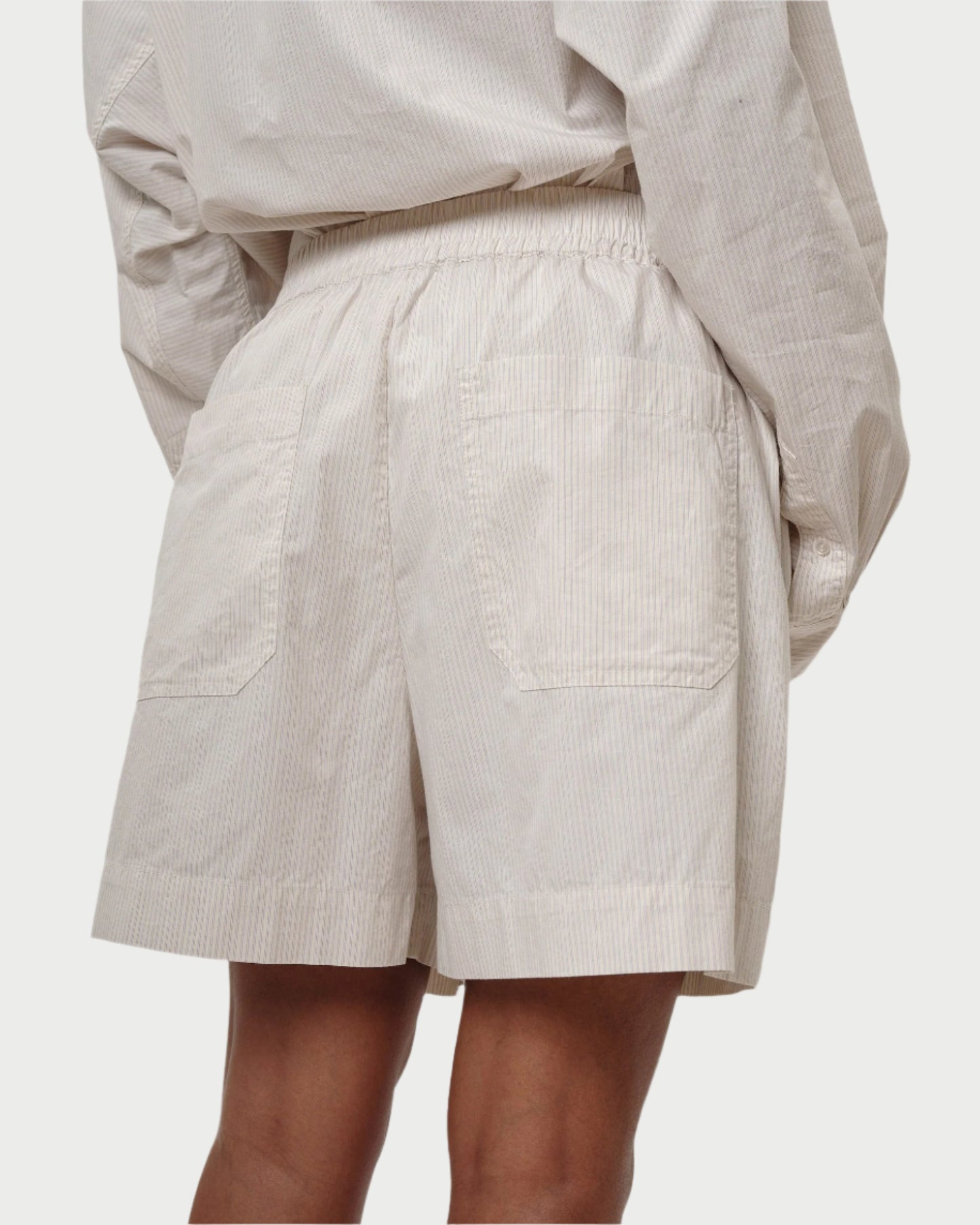 A person is wearing Herskind's Alma Shorts, featuring a relaxed fit, elastic waistband, and two back pockets. Paired with a loose-fitting, striped shirt made of 100% cotton with a subtle pattern, this ensemble is comfortable and casual.