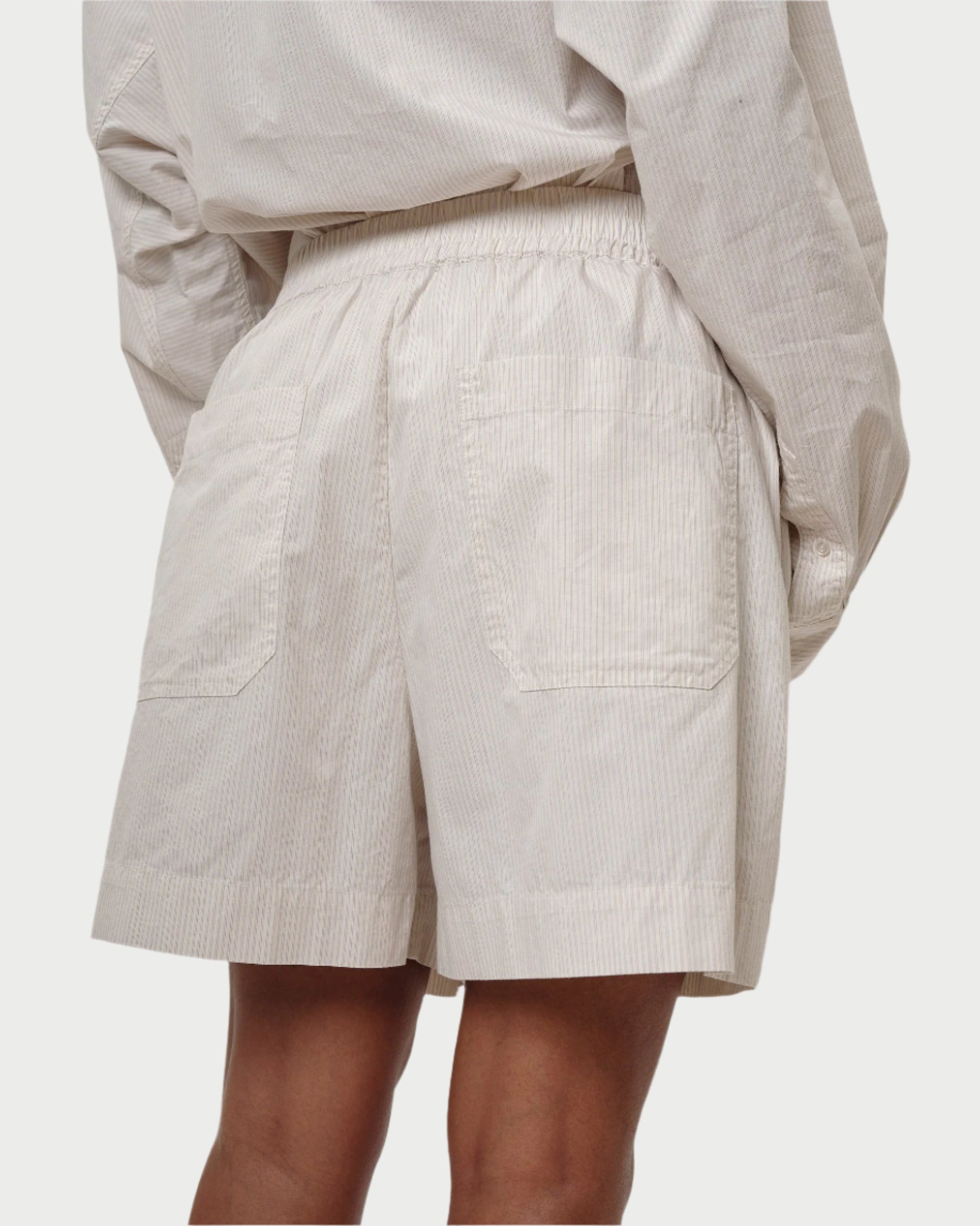 A person is wearing Herskind's Alma Shorts, featuring a relaxed fit, elastic waistband, and two back pockets. Paired with a loose-fitting, striped shirt made of 100% cotton with a subtle pattern, this ensemble is comfortable and casual.