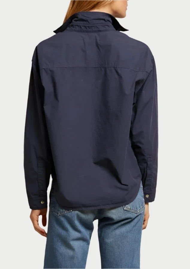A person with long blonde hair is standing with their back to the camera, wearing a dark blue James Button Down Shirt by perfectwhitetee and light blue jeans. The background is a plain, light-colored wall, creating an effortlessly stylish scene.