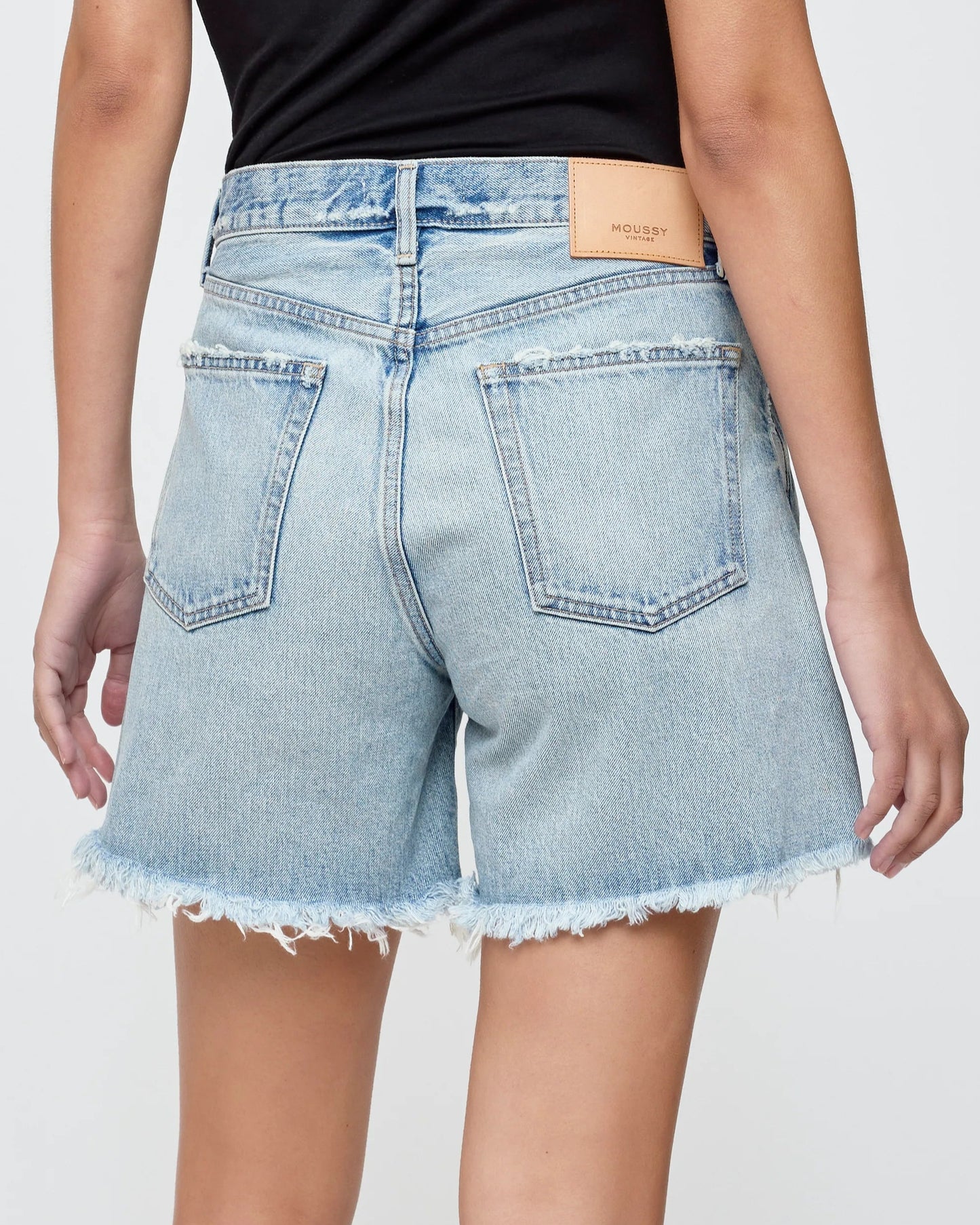 From the back, a person wears Moussy's Nolita Denim Shorts—light blue, high-waisted cotton denim with frayed hems and a tan leather waistband patch—paired with a simple black top for a perfect summer look.
