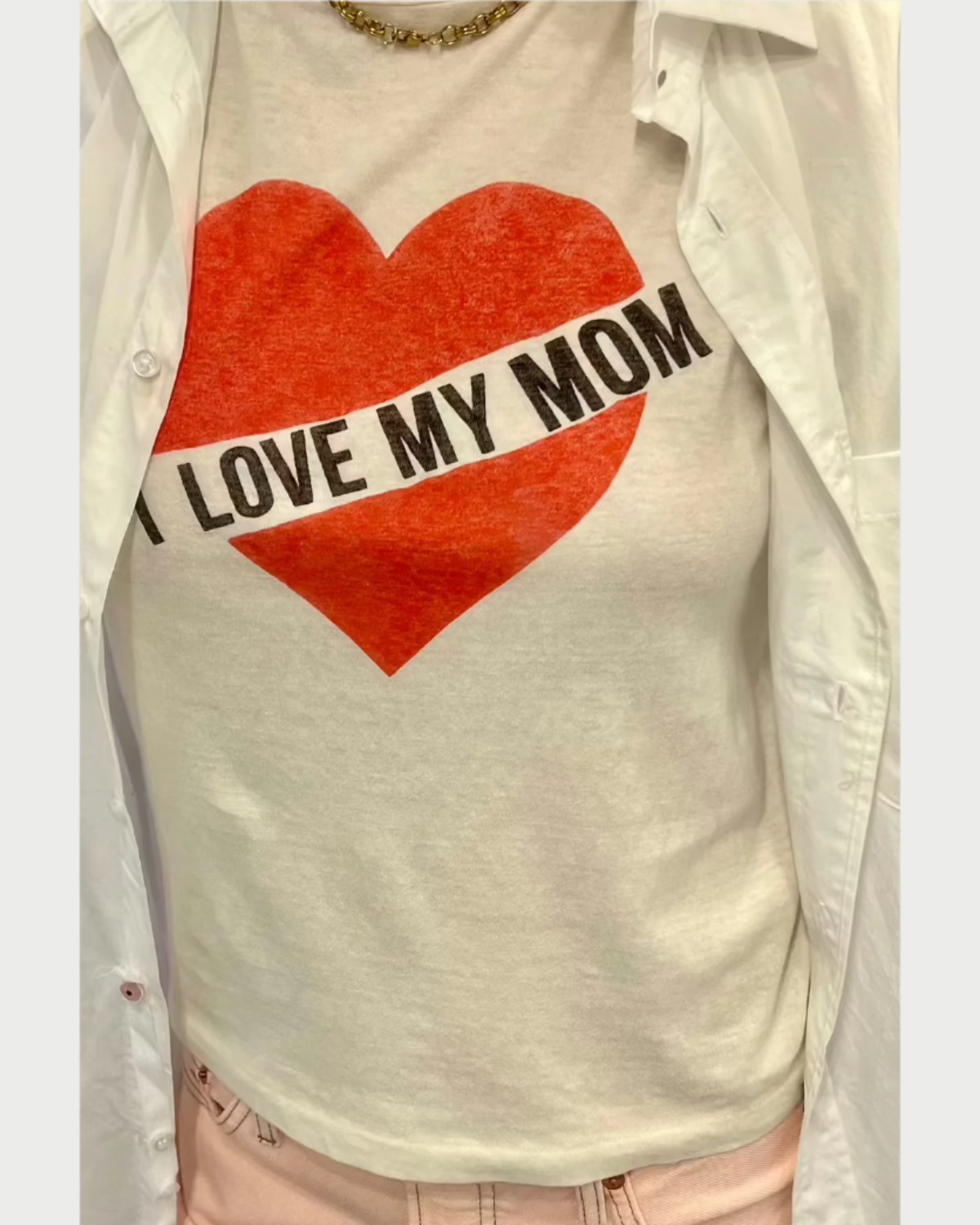 The individual is wearing a snug I Love My Mom T-Shirt by RE/DONE, crafted from 100% cotton and adorned with a red heart and the phrase "I LOVE MY MOM." It’s stylishly layered under an open white jacket and matched with fashionable light pink pants.