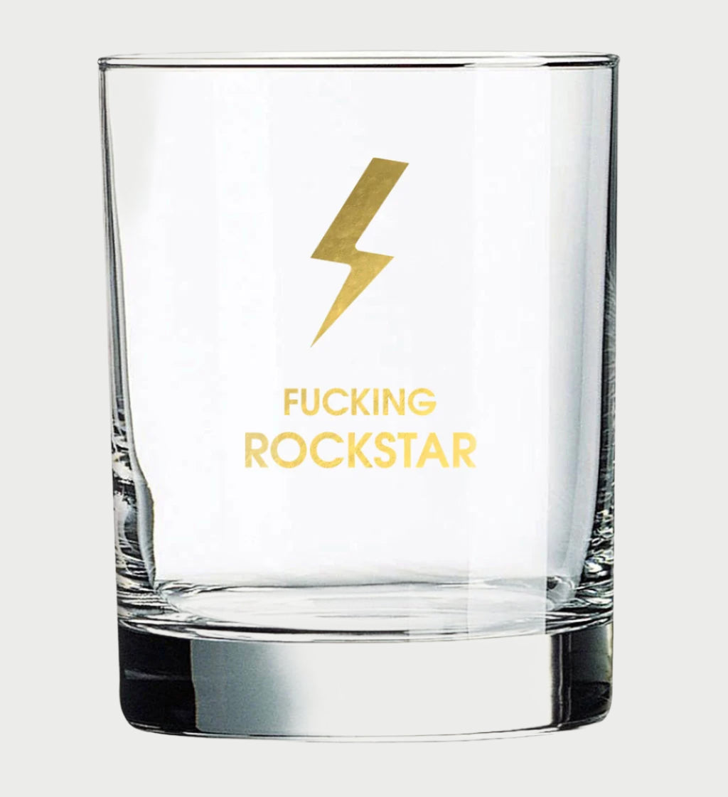 The More Rocks Glasses by Chez Gagné showcase a clear cocktail glass featuring a gold lightning bolt and bold "FUCKING ROCKSTAR" in gold foil, set brilliantly against a plain backdrop.