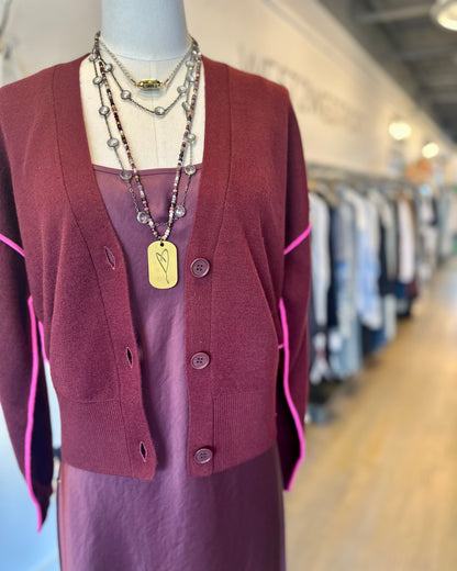 A mannequin in the Ileo Cardigan by Essentiel Antwerp, a mid-weight burgundy knit with contrast piping, layered over a silky dark mauve dress and necklaces, stands in a boutique with blurred racks behind.