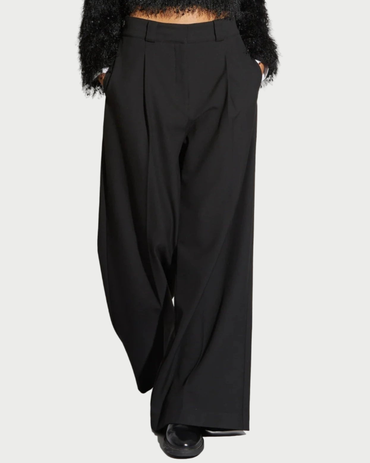 A person wears Saint Art NY's Tiffany Wide Leg Pants in black, a textured black sweater, and black shoes, standing with hands in pockets against a plain background.