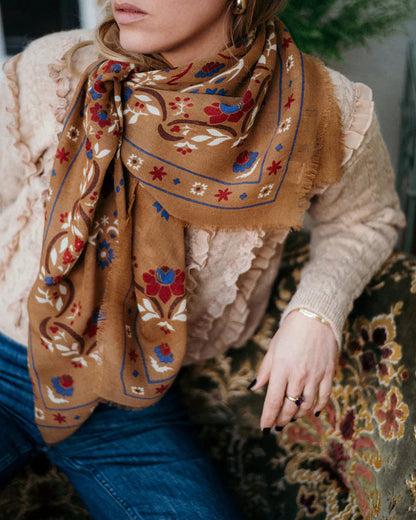 A woman in a textured beige sweater and blue jeans wears the Maison Lecomte Flament Secret Garden Scarf—an elegant wool and silk accessory by Maison Lecomte Flament with red, blue, and white floral patterns—while sitting on a patterned armchair.