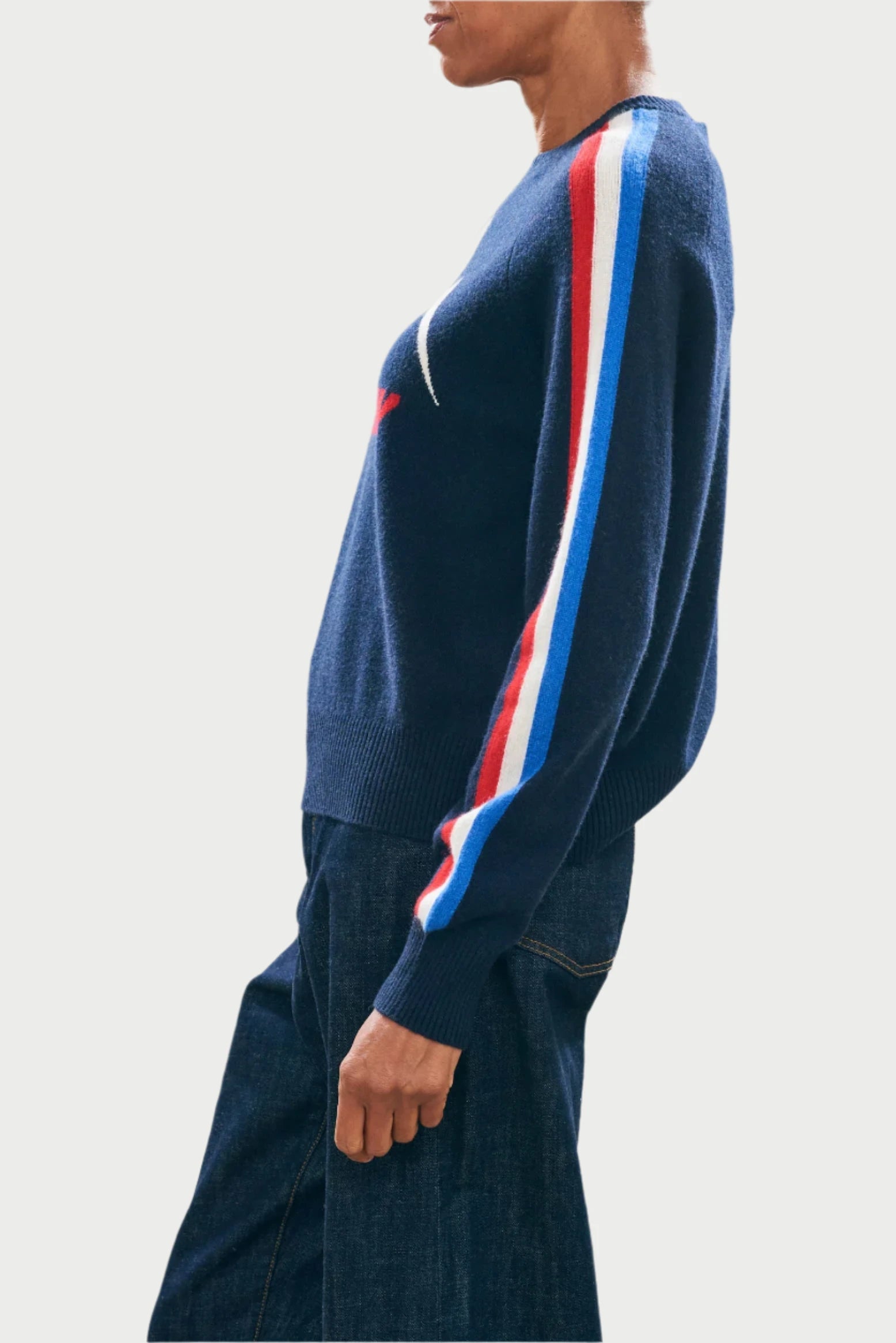 A person is standing in profile view against a light background, wearing the Ski Sunday Sweater by JUMPER, which is a navy piece with red, white, and blue stripe details on the sleeves, paired with dark jeans.