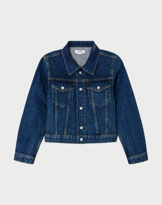 The RE/DONE Baby Trucker Jacket is a classic shrunken design in rich indigo Italian denim, featuring long sleeves, a button-up front, two chest pockets with button flaps, and a slightly faded texture reminiscent of a 70’s jean jacket. Displayed against a plain white background.