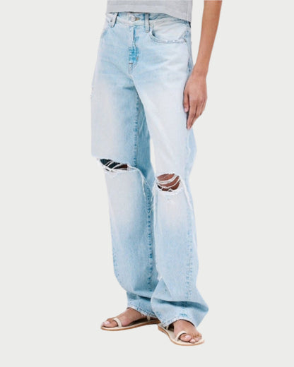 A person wearing a light gray t-shirt and SLVRLAKE's Tess Long Jeans, styled in a vintage-inspired low-rise fit with light blue fabric and knee tears, stands in sandals against a plain white backdrop.