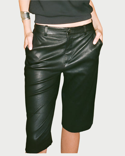 A person showcases SPRWMN's sleek Bermuda Trouser Shorts, crafted from supple French stretch-leather, with hands in pockets. A silver bracelet adorns their left wrist against a plain light green background.