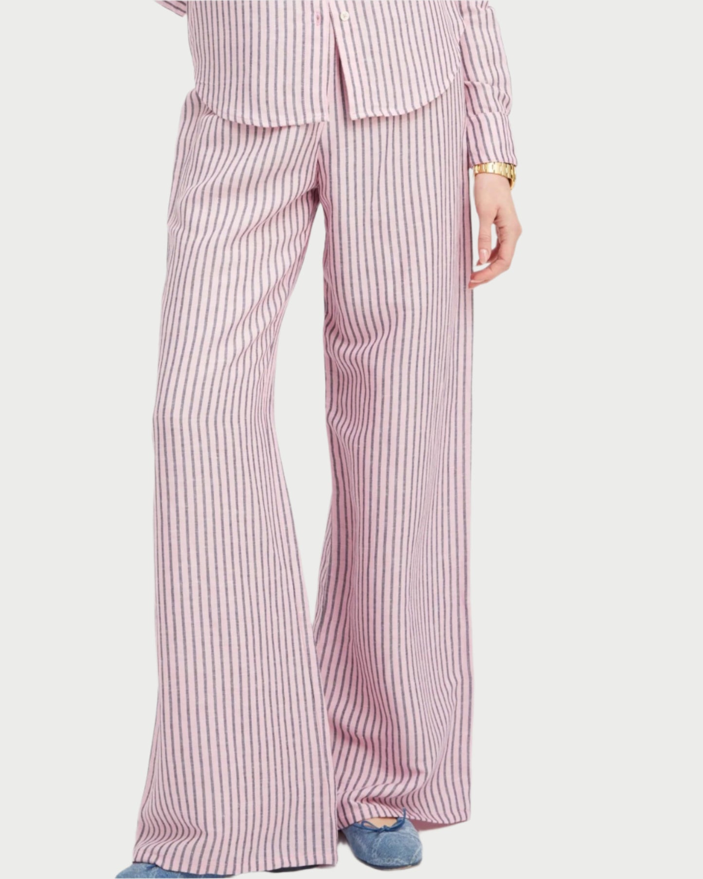A person, shown from the chest down, wears SPRWMN's Wide Leg Stripe Pants in pink and white stripes with an elastic waistband, a matching top, and blue slippers.