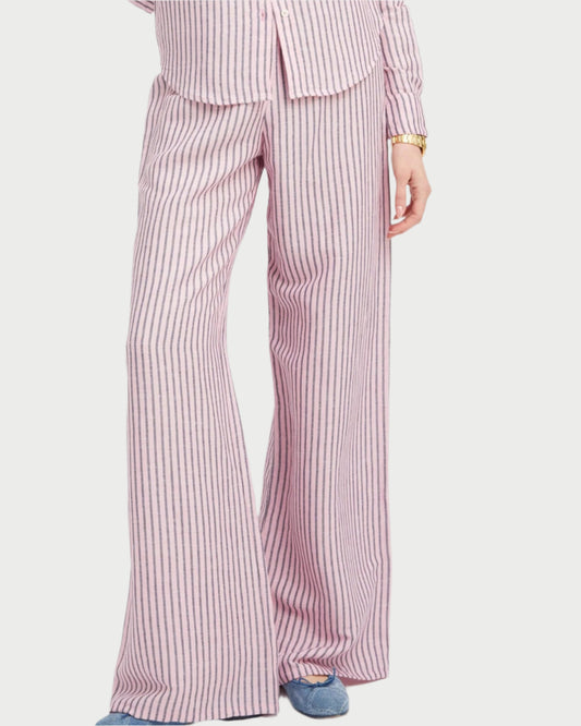 A person, shown from the chest down, wears SPRWMN's Wide Leg Stripe Pants in pink and white stripes with an elastic waistband, a matching top, and blue slippers.