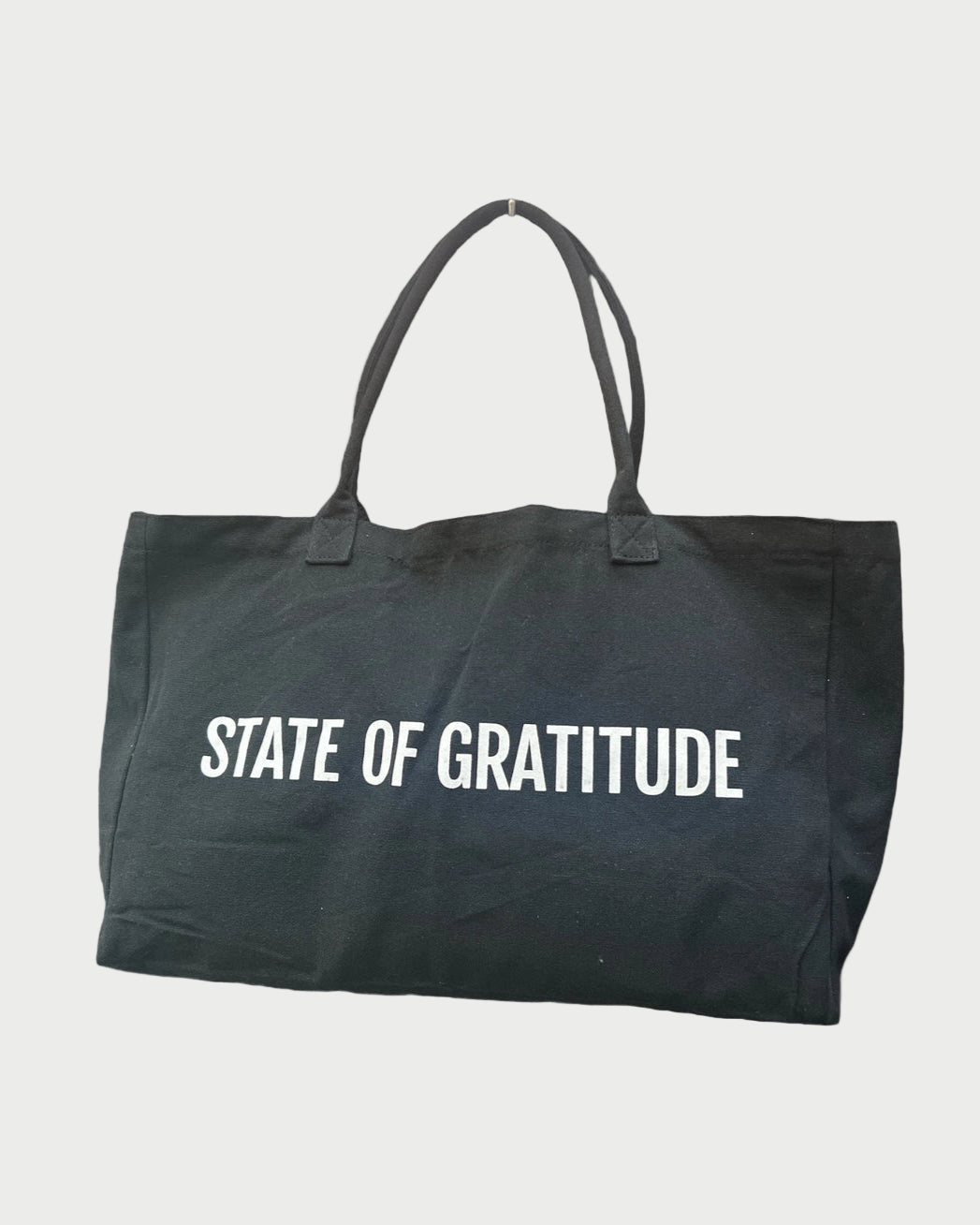 The State Of Gratitude Tote by State Of Gratitude is a large black organic bag with "STATE OF GRATITUDE" in bold white letters. Featuring two handles, it’s ideal for the beach or yoga and is shown on a plain white background.