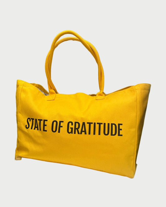 The State Of Gratitude Tote by State Of Gratitude is a spacious yellow organic bag with two handles and "STATE OF GRATITUDE" boldly printed on the front—ideal for carrying essentials to yoga or the beach.