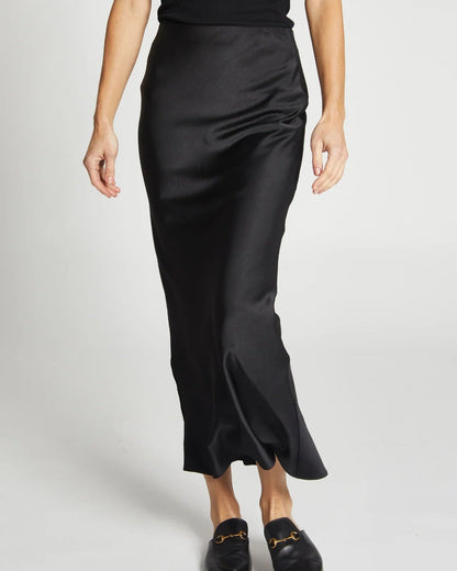 A person wearing the Saint Art NY Charmeuse Maxi Skirt in Black, a sleeveless top, and black loafers stands against a plain light background. The image shows the Saint Art NY skirt outfit from shoulders down.