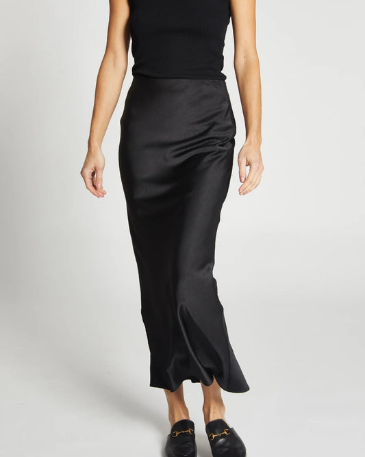A woman in a sleeveless black top, Charmeuse Maxi Skirt in Black by Saint Art NY, and black loafers stands against a plain white background.