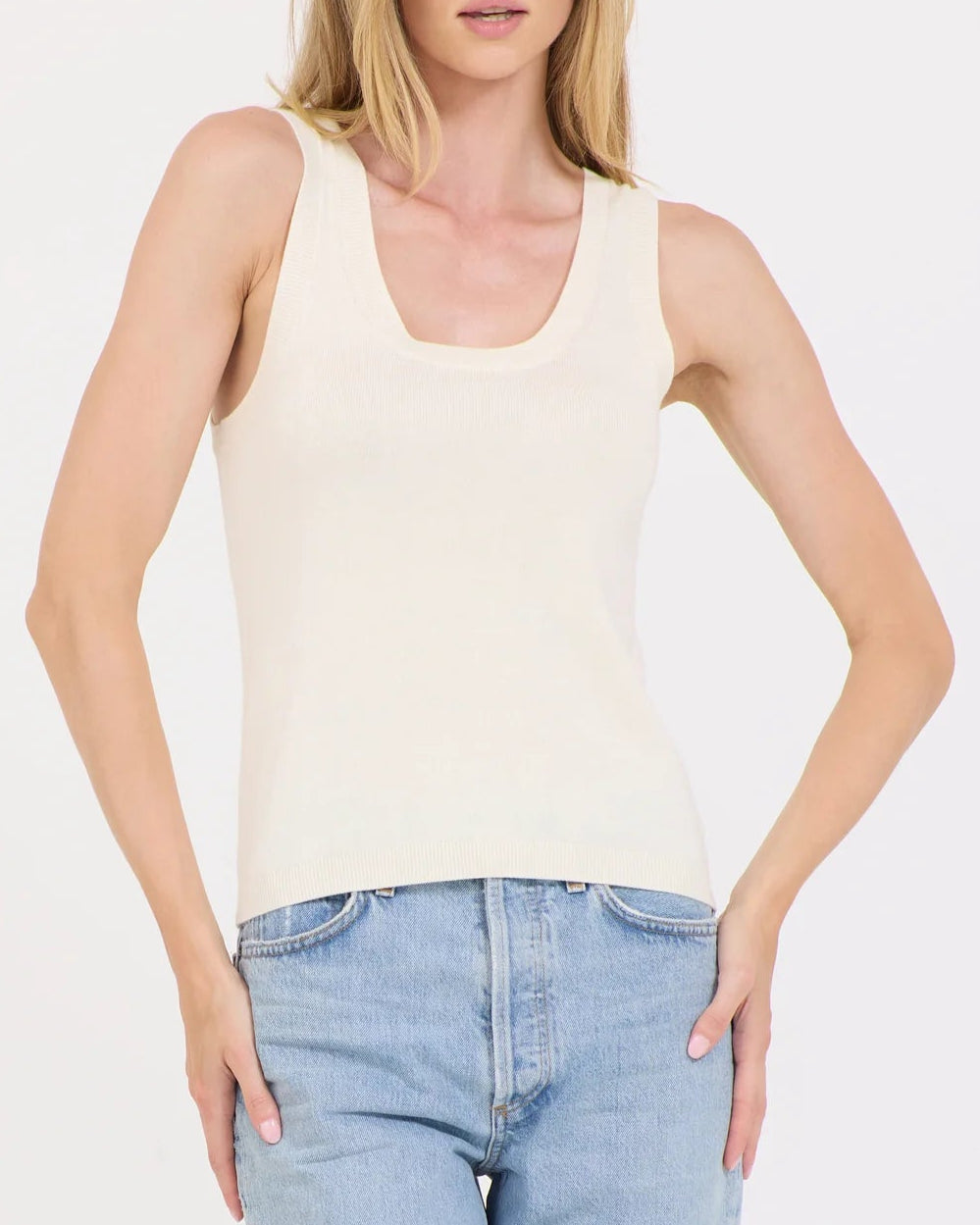 A person models the Terra Tank by One Grey Day, featuring a cream-colored, lightweight cotton design with a scoop neckline and paired with blue jeans. They stand against a simple background with their hands on their hips.