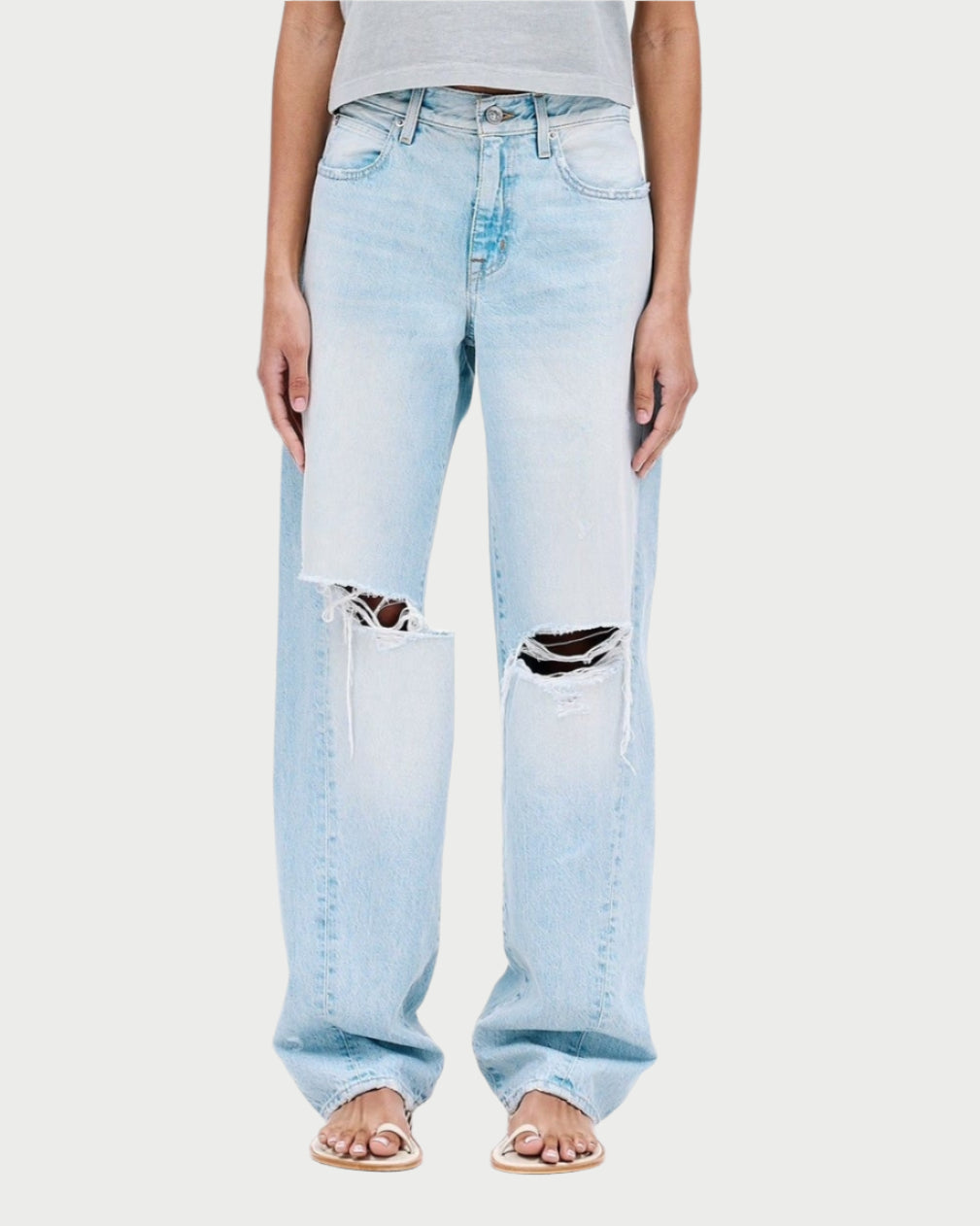 A person models a gray short-sleeve top with SLVRLAKE's Tess Long Jeans in light blue. These high-waisted, distressed jeans have knee rips for a vintage-inspired finish. White sandals complete the look against a plain background, emphasizing the stylish outfit.