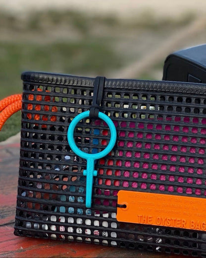 The Clutch by THE OYSTER BAG, a black mesh waterproof beach bag with an orange handle and label, rests on wood. A turquoise key-shaped keychain is attached. Lush greenery is blurred in the background, making this your perfect summer accessory.
