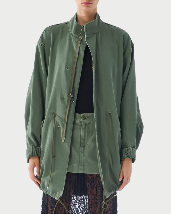Someone is wearing an olive green Utility Anorak from Le Superbe with a high collar and button details over a black top and skirt. The jacket has drawstrings and layers beautifully over an olive green lace-hem skirt. The background is plain.