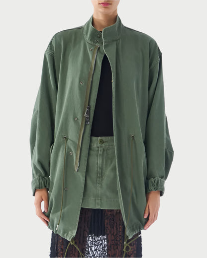 Someone is wearing an olive green Utility Anorak from Le Superbe with a high collar and button details over a black top and skirt. The jacket has drawstrings and layers beautifully over an olive green lace-hem skirt. The background is plain.