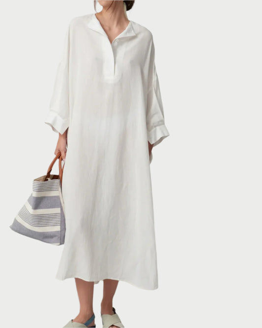 A person stands against a white brick wall wearing the WE-AR4 Oversized Kaftan, styled as a shirt dress with rolled sleeves, paired with a large striped tote bag and slip-on shoes.