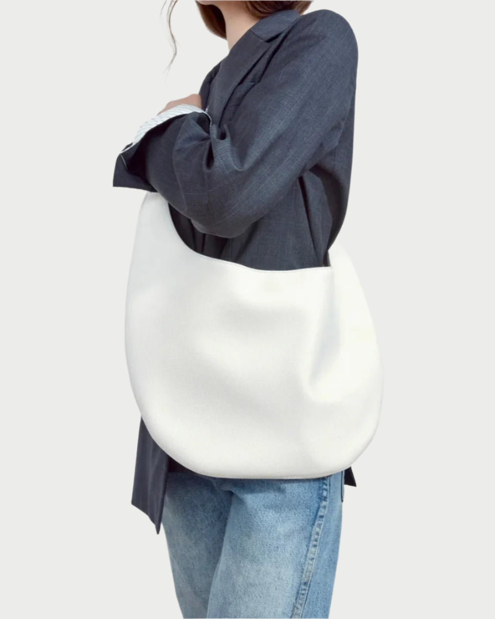 A person in a dark blazer and light blue jeans holds The H Hobo Bag by WE-AR4, crafted from upcycled grain leather, against a light gray background.