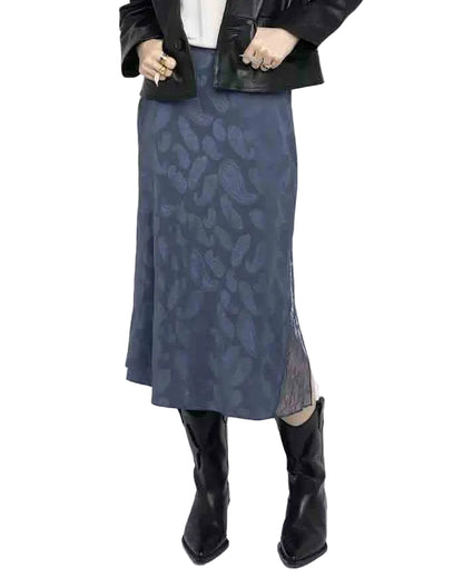 A person wears a black jacket, white top, Zadig & Voltaire Jayla Paisley Skirt—a blue paisley-print midi skirt with a side slit—and black pointed-toe boots, with their hands clasped at the waist.