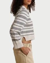 A woman stands in profile wearing a cropped long-sleeve striped knit top with navy and white horizontal stripes, paired with beige wide-leg pants and brown sandals, posed against a draped cream fabric backdrop.