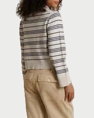 A model stands in profile wearing a cream and navy striped long-sleeve top and beige wide-leg trousers, posed against a draped cream fabric backdrop.
