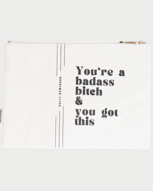 The Canvas Pouches by Properly Improper feature a durable white design with bold black text: "You're a badass bitch & you got this." Thin vertical lines and "DAILY REMINDER." accent the left, and a gold satin interior adds luxe flair.