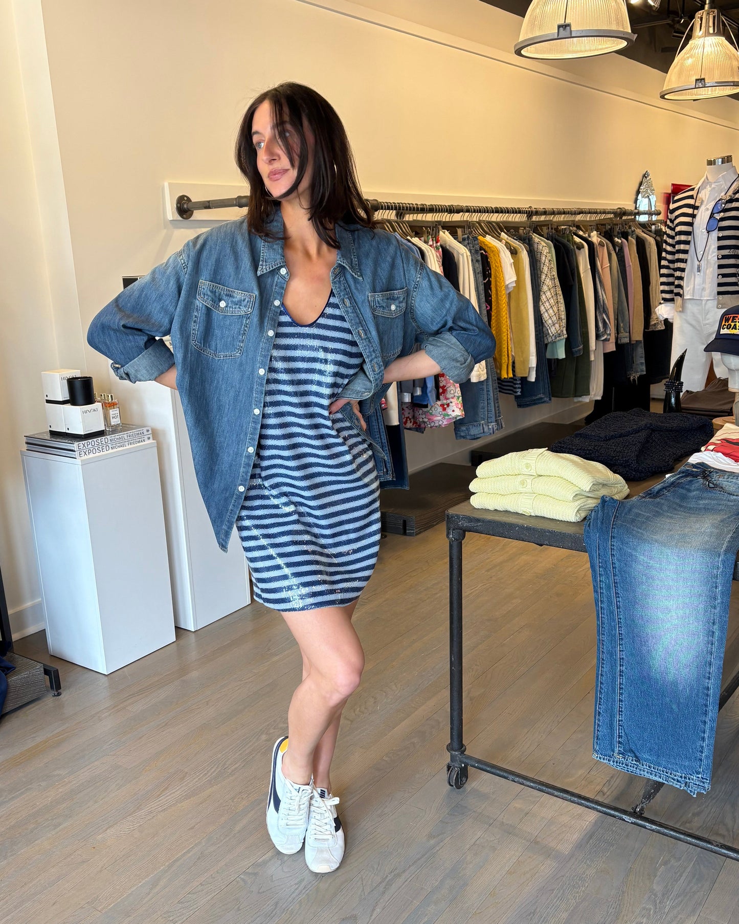 A woman poses in a clothing store, wearing the Zadig & Voltaire Rasha Sequin Stripe Dress, denim jacket, and white sneakers. She stands with one hand on her hip as French style dresses fill the racks and tables around her.