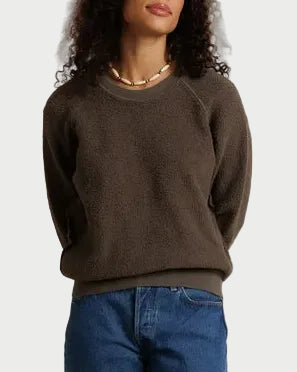 A woman stands against a draped off-white fabric backdrop wearing a dark brown crewneck sweater, blue jeans, and dark brown shoes.