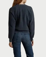 A woman stands with her back to the camera wearing a dark navy crewneck sweatshirt and blue flared jeans, standing against a draped beige fabric backdrop.