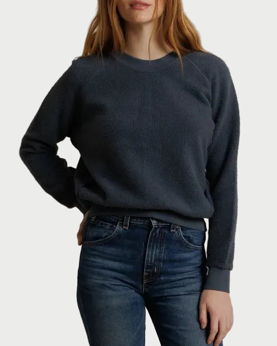 A woman with red hair wearing a dark slate blue crewneck sweatshirt and dark wash blue jeans, standing against a draped beige fabric backdrop.