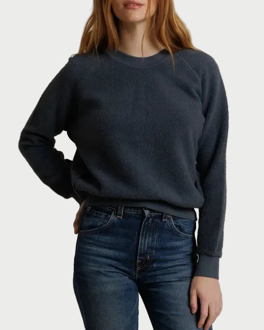 A woman with red hair wearing a dark slate blue crewneck sweatshirt and dark wash blue jeans, standing against a draped beige fabric backdrop.